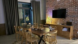A dining area with a wooden table and chairs, a TV mounted on a brick wall, and a window with curtains. - MyGarden Villa Binh Chau Hotspring (Binh Chau)