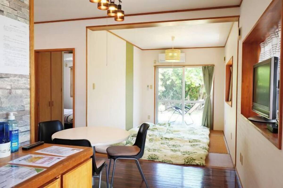 Ein Zimmer mit einem Holztisch, Stühlen, einem Bett mit floralem Überwurf, einem Fernseher und einem Fenster mit Blick auf Grünfläche.