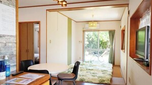 Una habitación con escritorio de madera, sillas, cama con colcha estampada y televisor, además de ventana con vista a follaje.