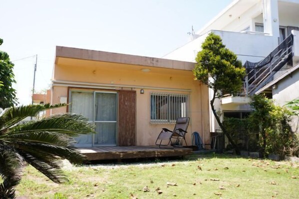 A modern two-story building with a flat roof, a balcony with a chair, and a small garden area.