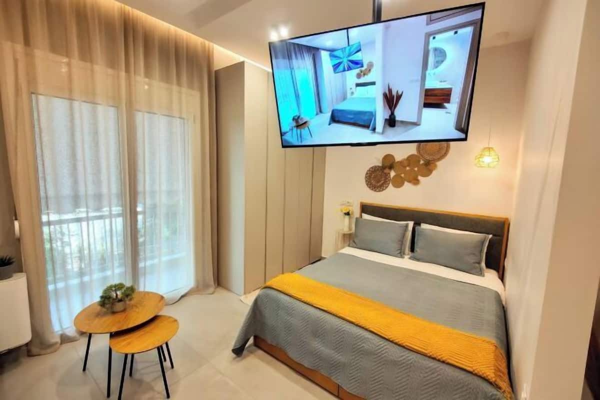 Ein modernes Hotelzimmer mit einem Bett, einem an der Wand befestigten Fernseher, einem kleinen Tisch mit Pflanze und einem großen Fenster mit durchscheinenden Vorhängen.