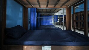 A room with a wooden ceiling, a bed with a dark mattress, and a view through a window with blue curtains. - Bunk Bed Bali near Sanur Mitra RedDoorz (Bali)