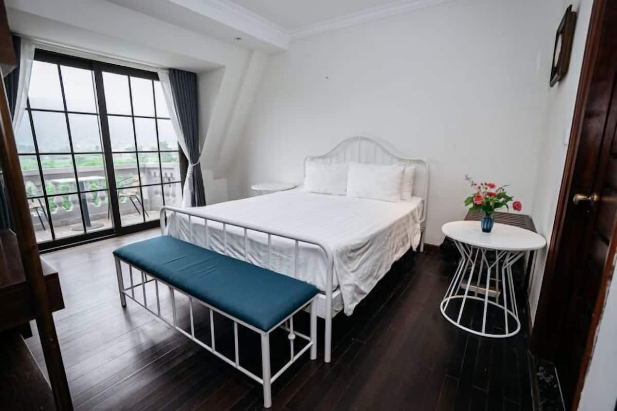 A bedroom with a bed, a bench, a small table with a vase of flowers, and a view through a window.