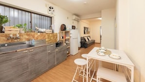 Una cocina moderna con una isla central, un comedor con mesa blanca y taburetes de bar, y una sala de estar con sofá y televisor.