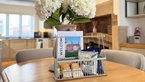 A welcome sign with a house illustration, a potted plant with white flowers, and a metal basket containing coffee bags and a bag of chips on a wooden table. - Gardener's Cottage (Lytham St. Anne's)