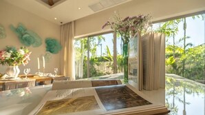 A room with a dining table set for two, a large window with a view of greenery, and a vase with flowers on the windowsill. - Villa Allamanda 3 Bdrm Best Location Bangtao Beach VVPHU88 (Phuket)
