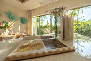 A room with a dining table set for two, a large window with a view of greenery, and a vase with flowers on the windowsill. - Villa Allamanda 3 Bdrm Best Location Bangtao Beach VVPHU88 (Phuket)