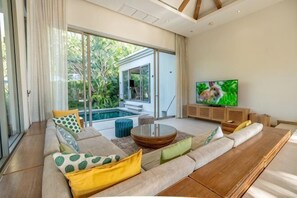 A modern living room with a large sofa, a coffee table, a TV, and a pool visible through the glass doors. - Villa Allamanda 3 Bdrm Best Location Bangtao Beach VVPHU88 (Phuket)