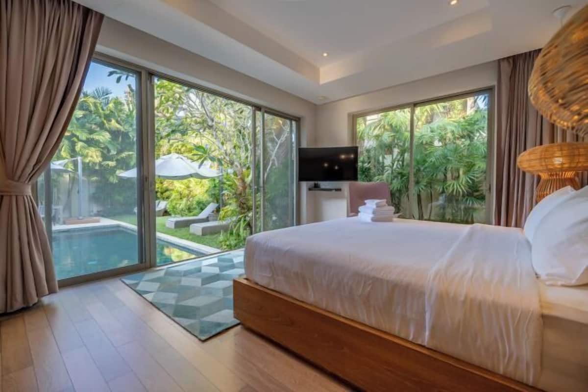 Ein Schlafzimmer mit einem großen Bett, einem Fernseher und Blick auf einen Swimmingpool und Grünflächen.