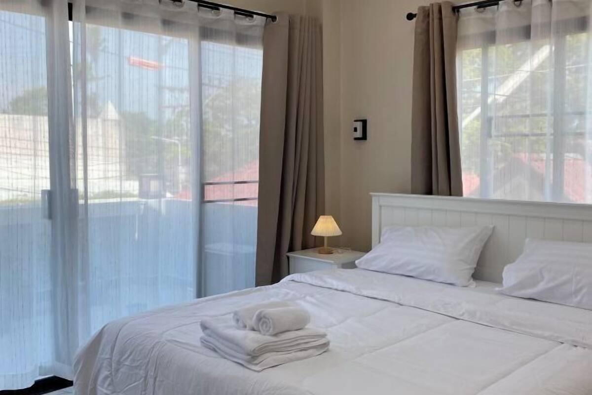 A hotel room with a bed, curtains, a nightstand with a lamp, and a view through the window.