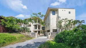 A multi-story residential building with a central entrance, surrounded by lush greenery and palm trees. - Amano Terrace (Onna)