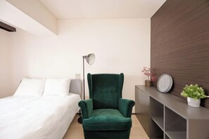 A modern hotel room with a green armchair, a bed with white linens, a bedside table with a clock and a potted plant, and a dark wood accent wall. - Shinjuku Kabukicho Harmony Inn (Tokyo)