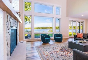 Uma sala de estar ampla, com janelas grandes, lareira e vista para um lago.