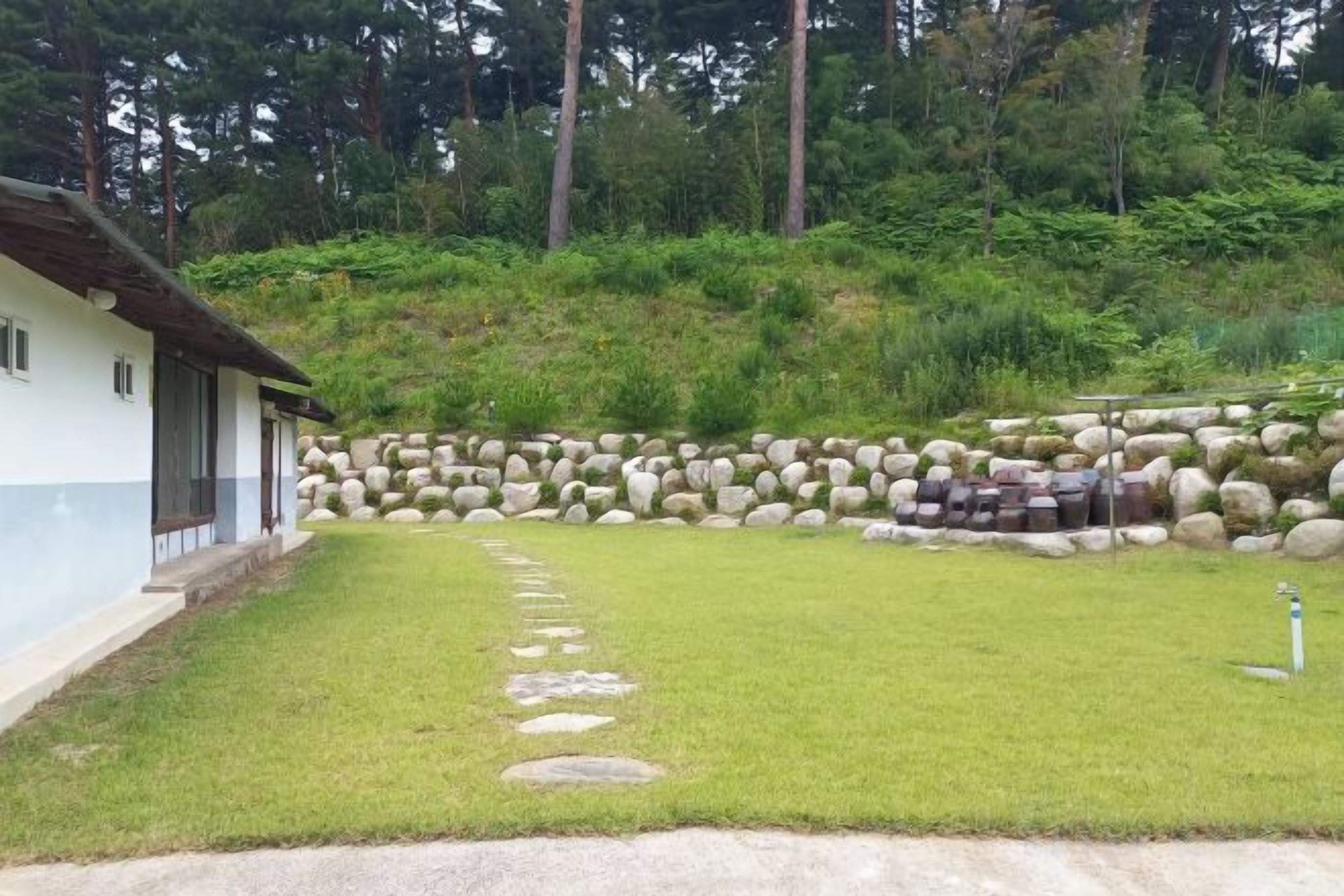 A house with a stone pathway leading to a white building, surrounded by a grassy area and a stone wall.