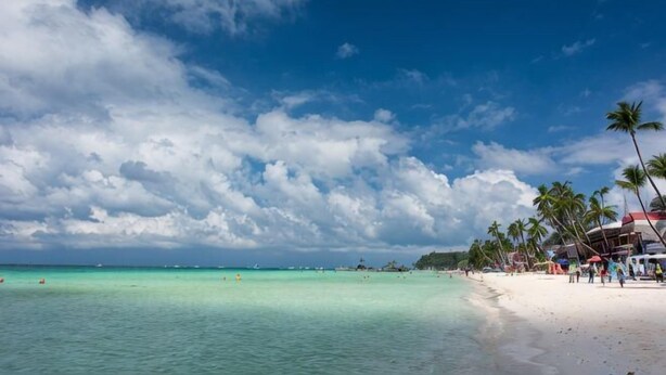 A beach with clear turquoise water, palm trees, and a few buildings in the background. - Super diving and tinas reef divers (Puerto Galera)
