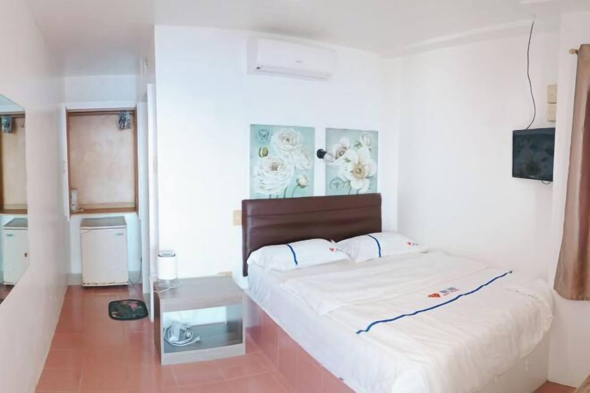 A bedroom with a bed, a small bedside table, a wall-mounted air conditioner, and a door leading to another room.