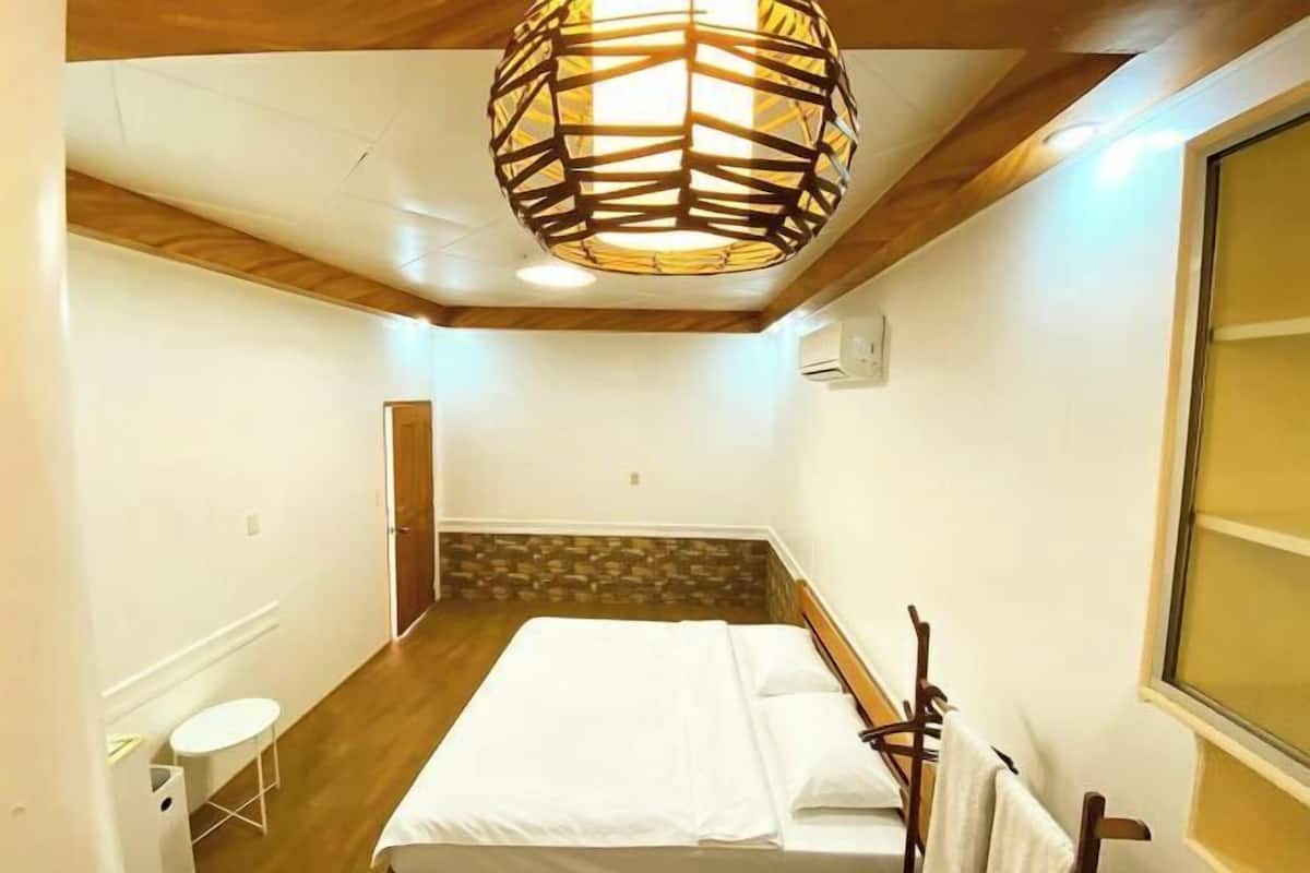 A hotel room with a bed, a small table, a hanging light fixture, and a window.