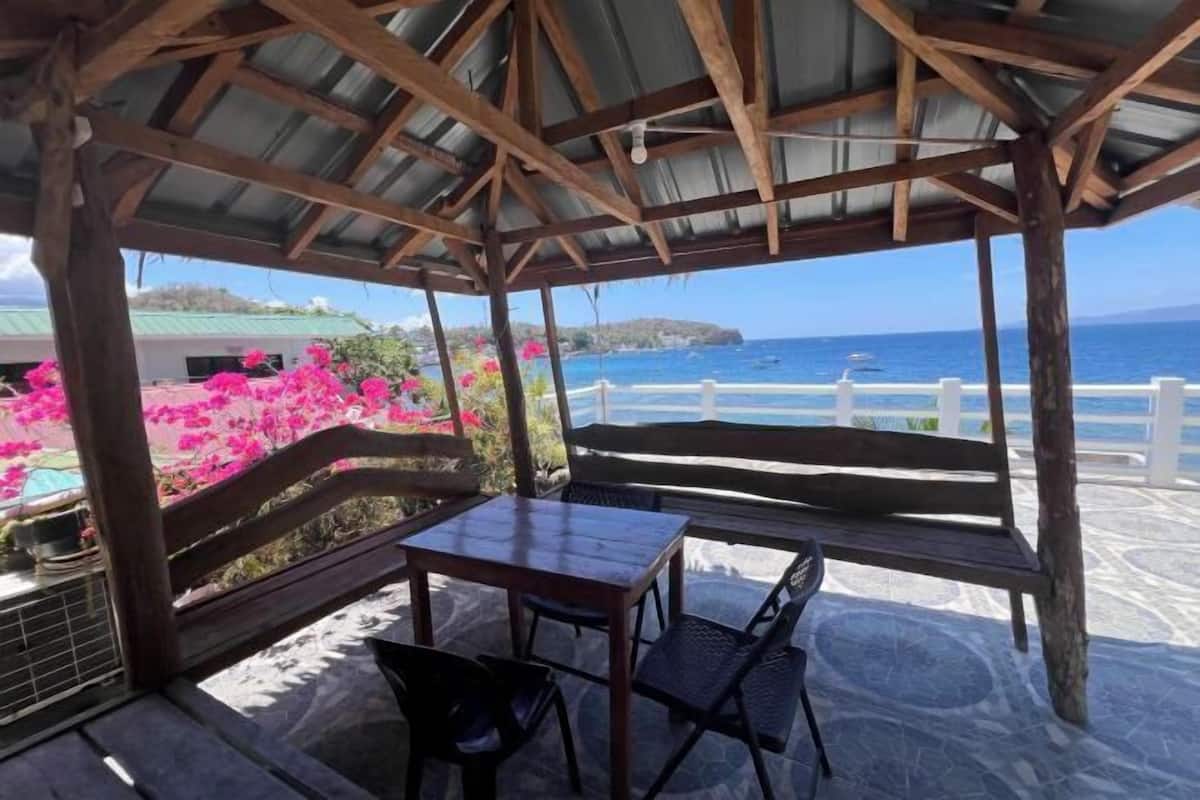 A covered outdoor seating area with a table and chairs, overlooking a beach and ocean.