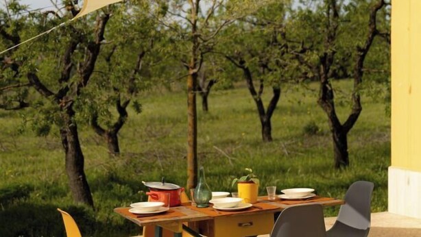 Un espace de restauration en plein air avec une table dressée pour deux personnes, entouré d’arbres et bordé d’un mur jaune.