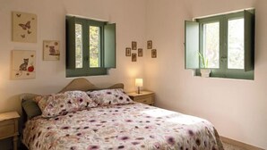Une chambre à coucher comprenant un lit, deux tables de chevet, une petite table, une lampe et deux fenêtres avec des volets verts.