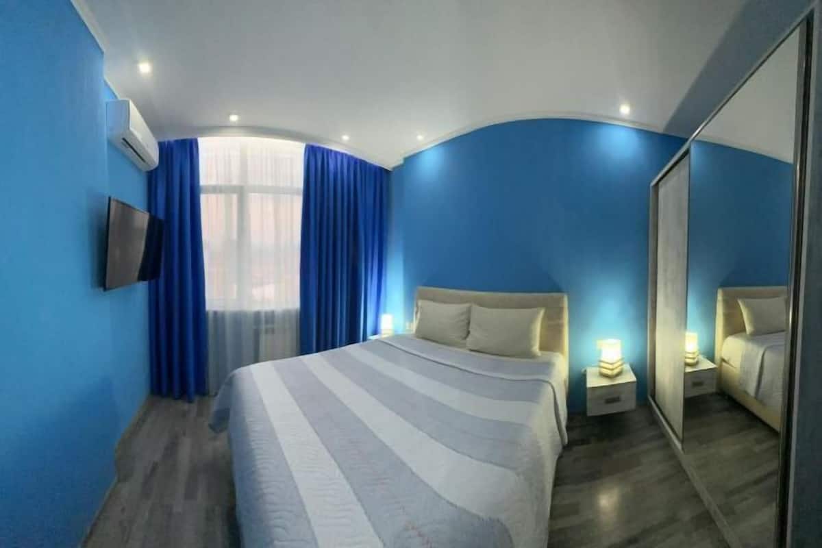 A hotel room with a large bed, a TV, a mirror, and a window with blue curtains.