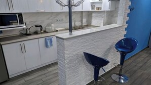 Une cuisine moderne avec des meubles blancs, un plan de travail en marbre, un évier et une machine à café. On y trouve deux tabourets de bar bleus et un mur bleu.