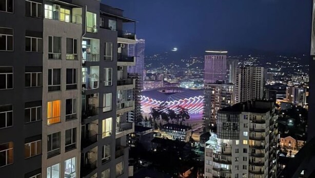 Une vue nocturne d’une ville, avec de hauts immeubles et une zone illuminée au loin.