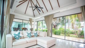 A modern living room with a sectional sofa, a ceiling fan, large windows, and a view of greenery outside. - Naya Poly Pool Villa-80 (Phuket)