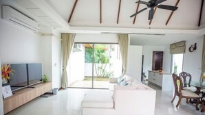 A modern living room with a white sofa, a TV on a wooden stand, and a dining area with wooden chairs. - Naya Poly Pool Villa-80 (Phuket)