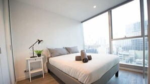 A modern bedroom with a large bed, a bedside table with a lamp, and a view of the city through large windows. - A Chic 2BR, CBD Apartment Near Southern Cross (Melbourne)