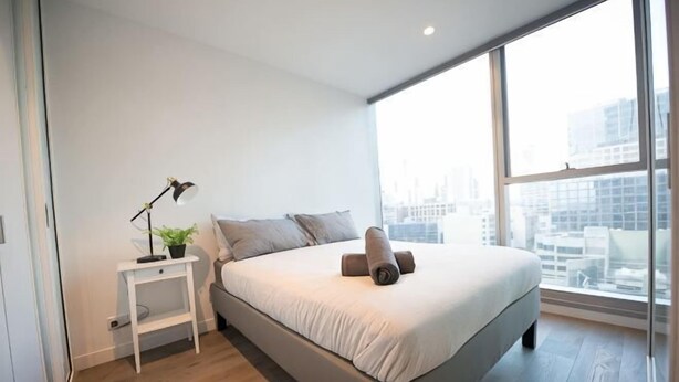 A modern bedroom with a large bed, a bedside table with a lamp, and a view of the city through large windows. - A Chic 2BR, CBD Apartment Near Southern Cross (Melbourne)