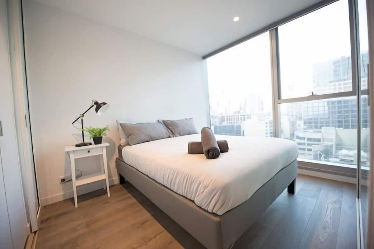 A modern bedroom with a large bed, a bedside table with a lamp, and a view of the city through large windows.