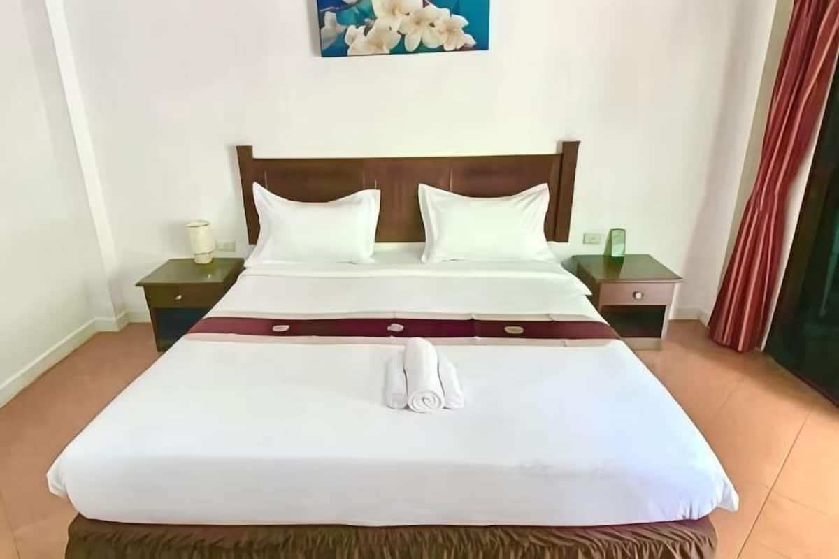 A neatly made bed with a white duvet and two pillows, a wooden headboard, a nightstand with a lamp, and a painting on the wall.