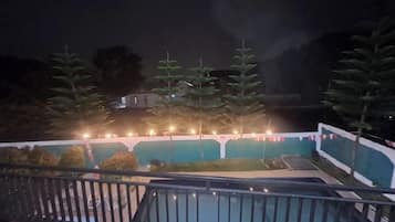 Vista nocturna de un balcón con barandilla, árboles y un edificio al fondo.