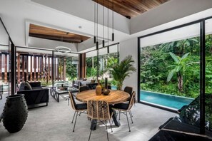 A modern dining area with a round wooden table, black upholstered chairs, and a large window overlooking a lush garden. - Villa Three Little Birds by BaliSuperHost (Bali)