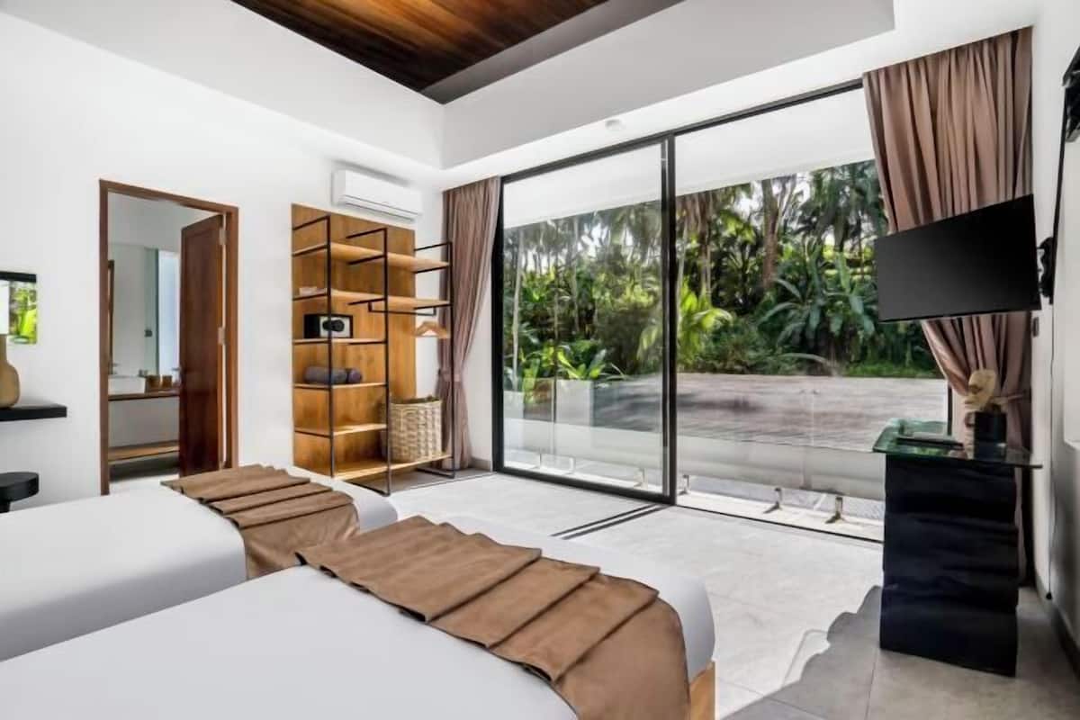 A modern bedroom with two beds, a large sliding glass door to a balcony, a TV, and a wooden shelving unit.