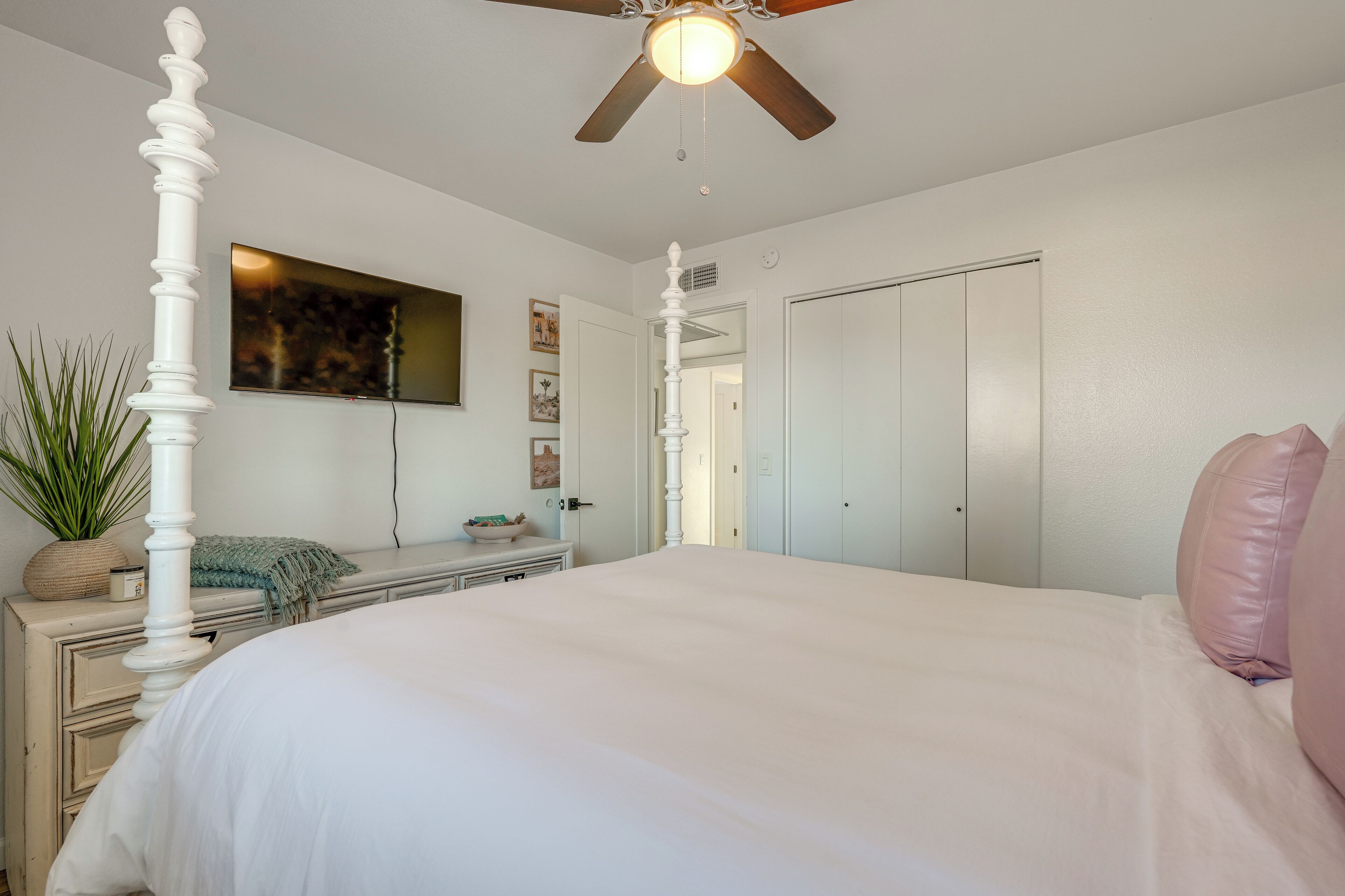 A bedroom with a four-poster bed, a ceiling fan, a dresser, a mirror, and a door leading to another room.
