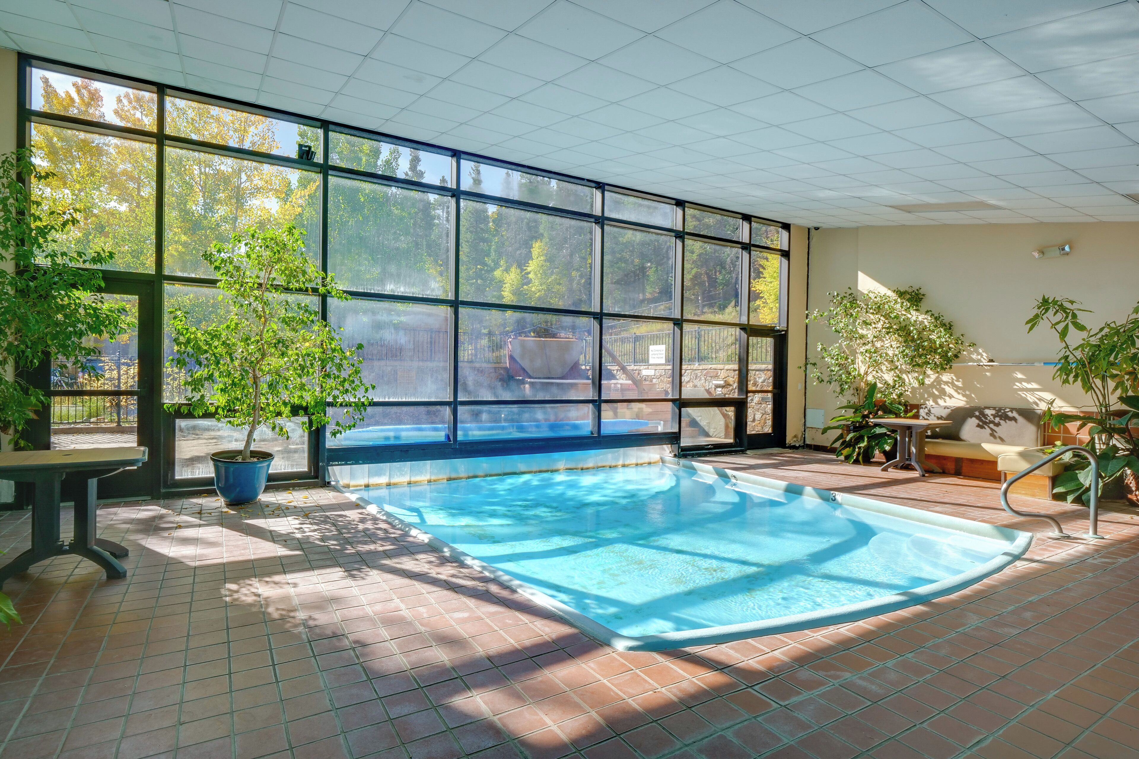 An indoor swimming pool with tiled flooring, large windows, and potted plants.