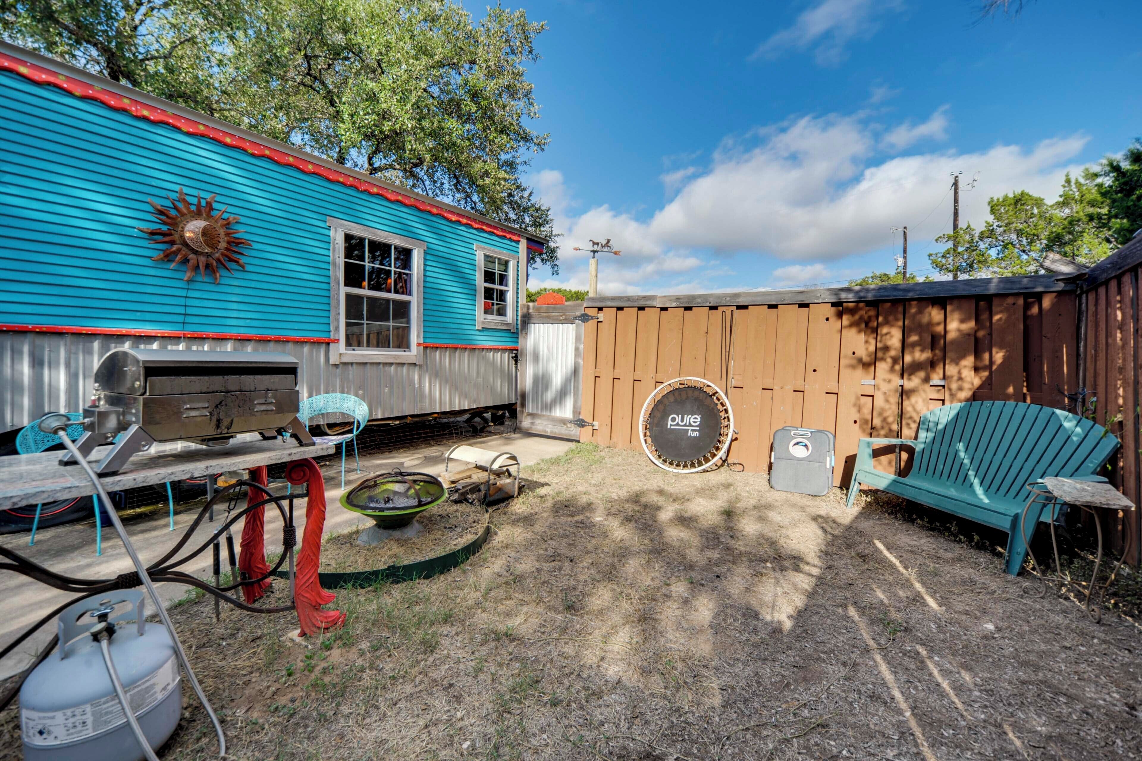 Un patio trasero con una casa azul, una cerca de madera, una parrilla, una silla azul y un paraguas rojo.