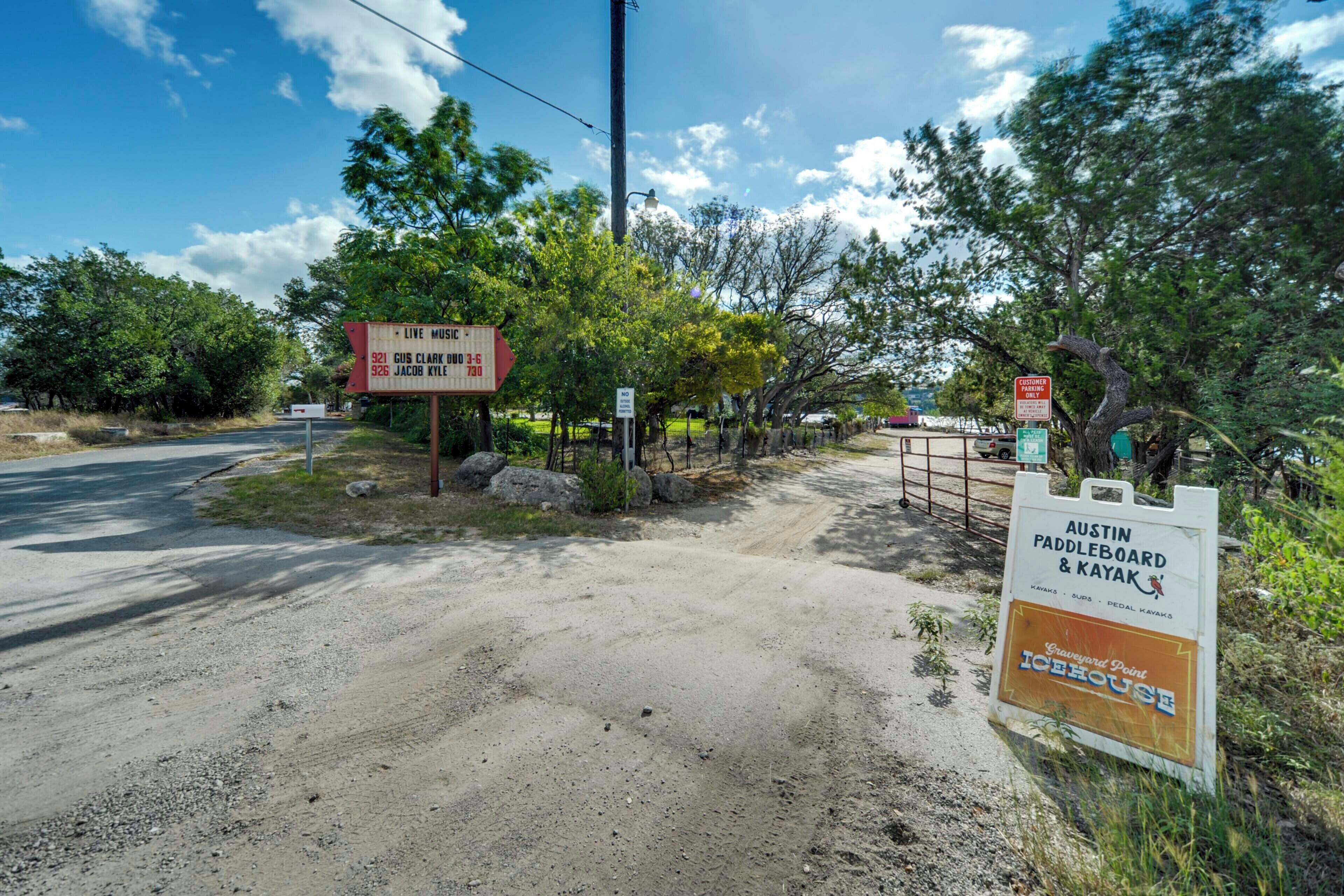 Un chemin de terre mène à une zone fermée par une barrière, où se trouvent des panneaux indiquant les lieux d’Austin Paddleboard & Kayak et les événements de musique en direct.