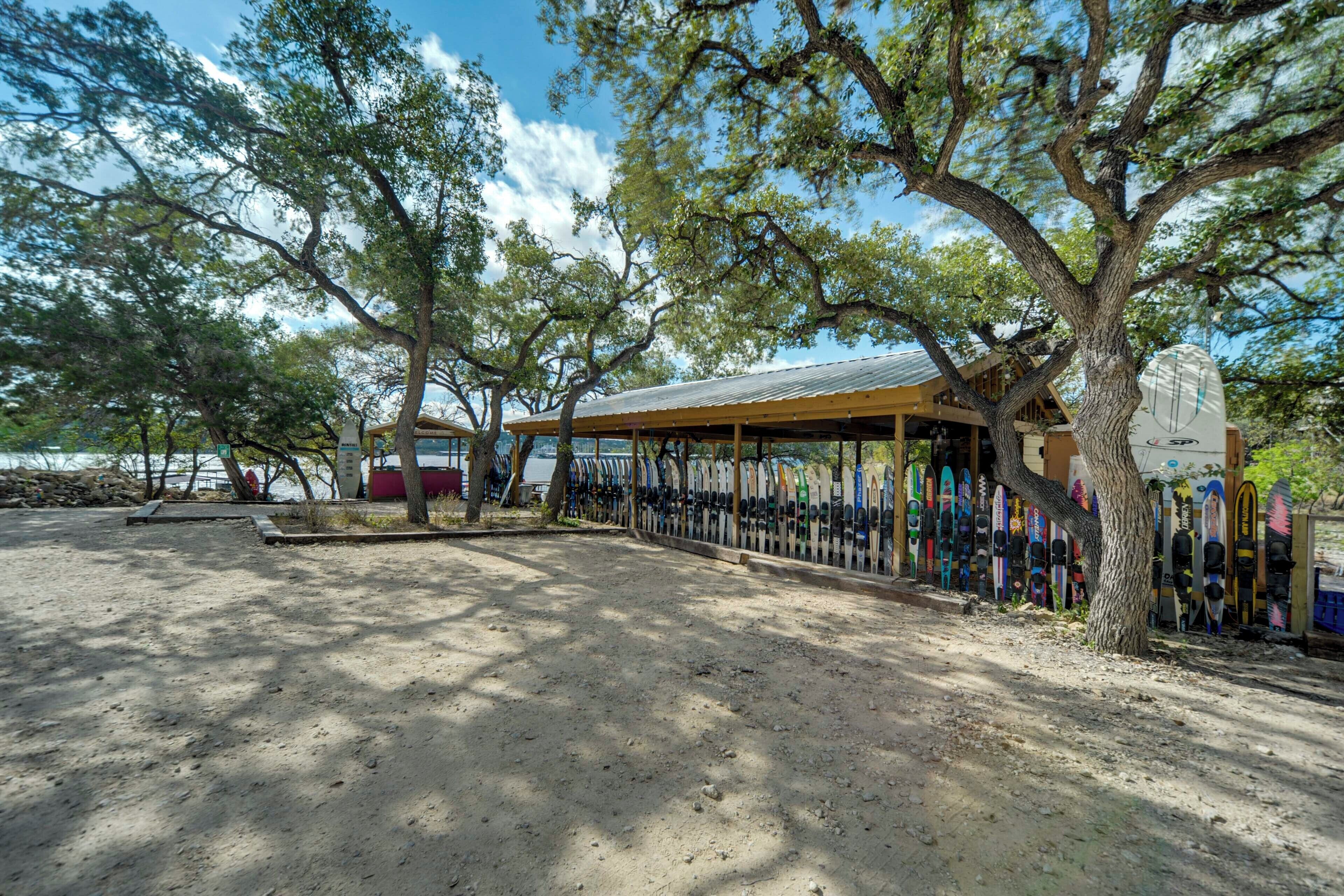 A covered outdoor area with a row of surfboards, a building with a metal roof, and trees providing shade.
