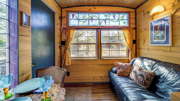 A cozy cabin interior with a wooden ceiling, a large window with curtains, a leather sofa with decorative pillows, and a dining table with glassware. - Walk to Lake Travis: Austin Tiny Home w/ Hot Tub! (Lakeway)
