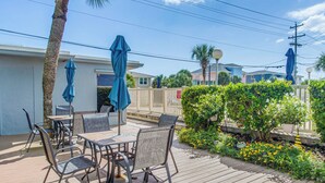 A wooden deck with outdoor furniture and umbrellas, surrounded by greenery and a white fence. - Steps to Shore + Pier: Condo in North Myrtle Beach (North Myrtle Beach)