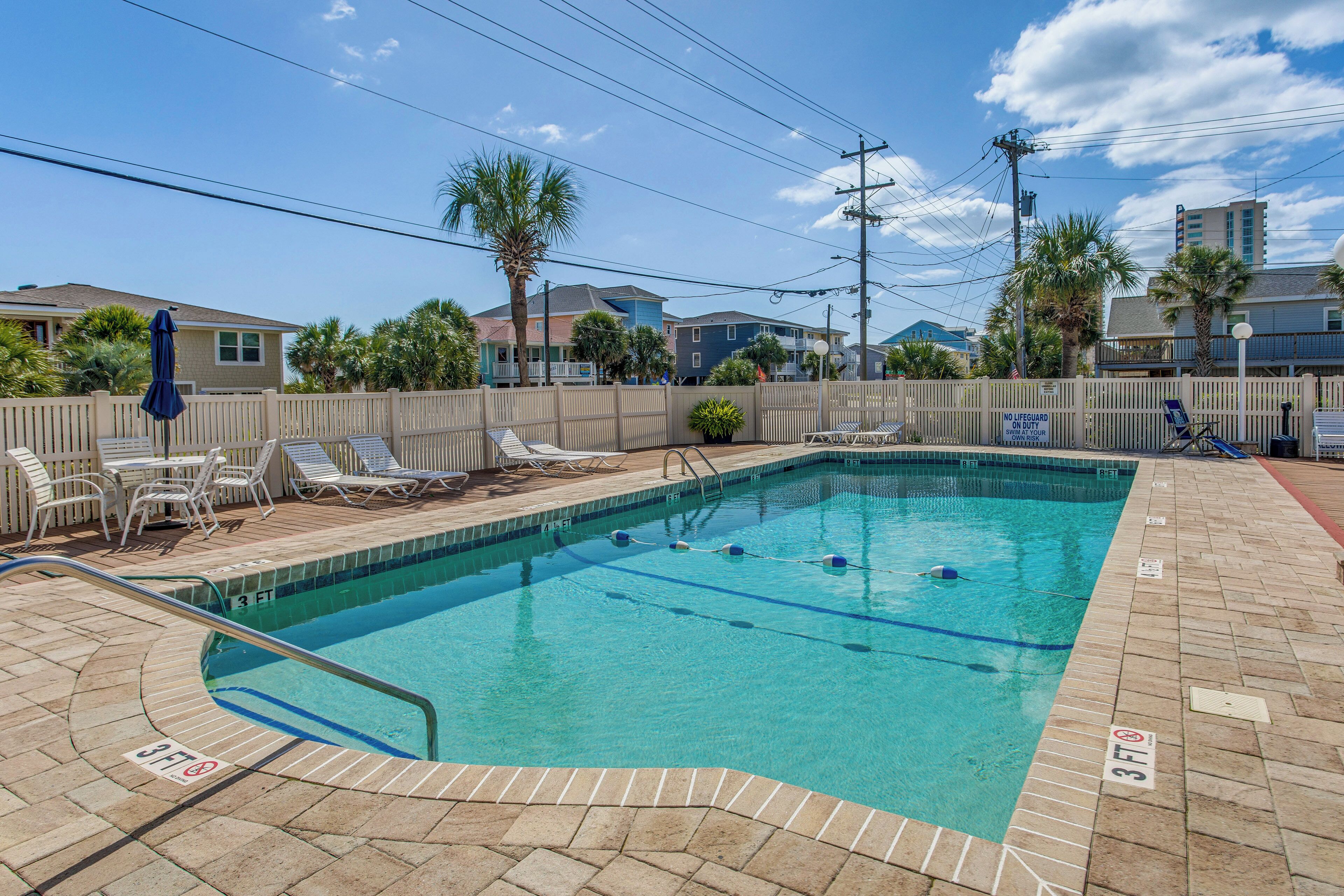 Una piscina rectangular con plataforma azulejada, rodeada por una cerca blanca, y un letrero que indica ausencia de salvavidas.