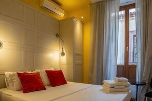 A hotel room with a bed, pillows, a lamp, a window with curtains, and an air conditioning unit. - Apartamento Moderno Junto a la Gran Via (Madrid)