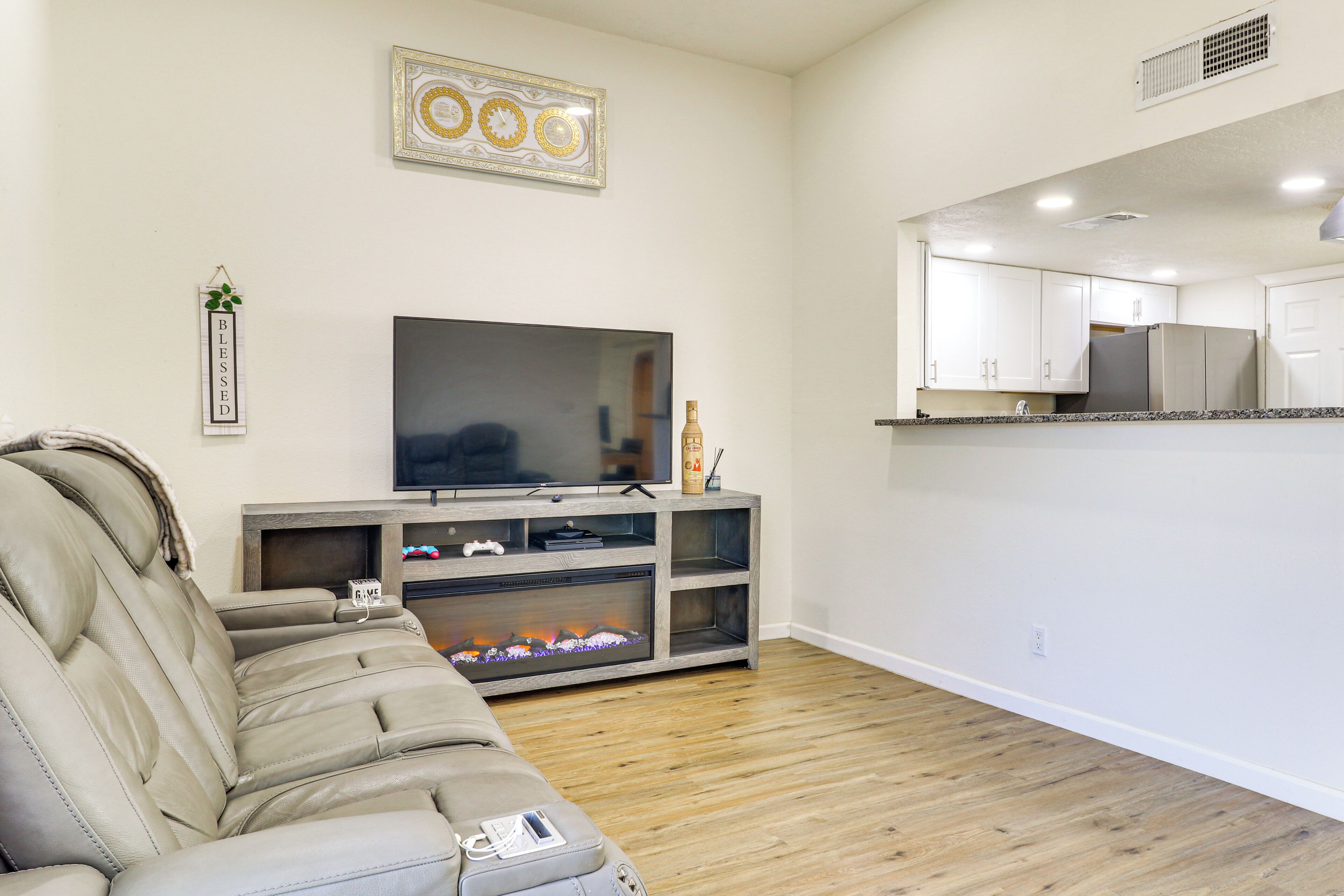 A living room with a leather sofa, a flat-screen TV, a fireplace, and a kitchen area with cabinets and a refrigerator.