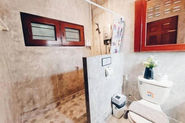 A bathroom with a toilet, a small trash bin, a shower area, and a window with a wooden frame. - The Warehouse - Ayutthaya (Ayutthaya)