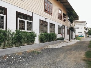 A white building with multiple windows, a gravel driveway, and a small garden area with shrubs. - The Warehouse - Ayutthaya (Ayutthaya)