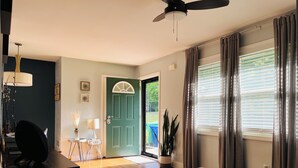 A spacious living room with a wooden floor, a ceiling fan, a door with a green door frame, and a view of the outdoors through large windows with blinds.