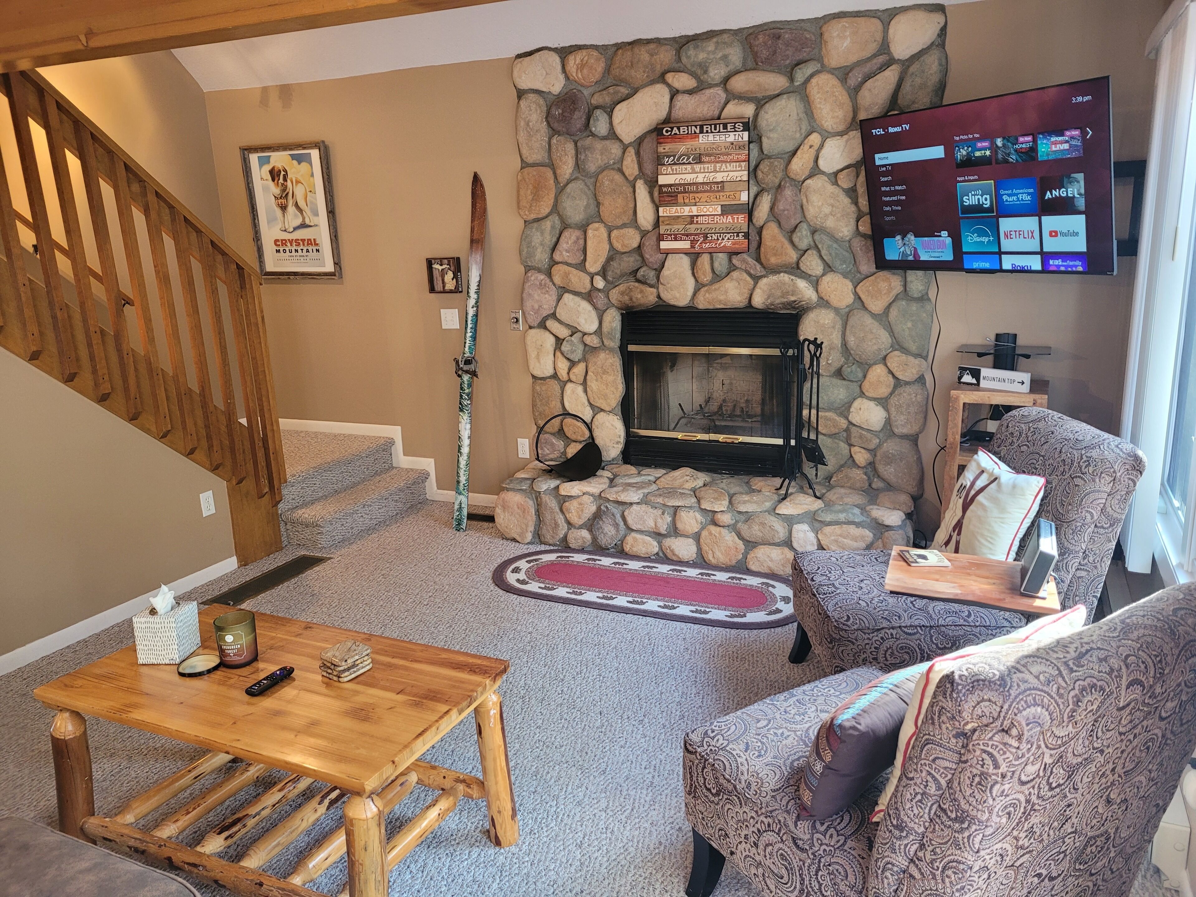 Una sala de estar acogedora con chimenea de piedra, televisor montado, escalera de madera y un cartel enmarcado.